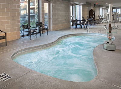 An indoor hot tub is visible, featuring a curved shape and soothing water. Surrounding seating is provided by several black chairs against the tiled wall. Large windows allow natural light to fill the space, enhancing the tranquil atmosphere.