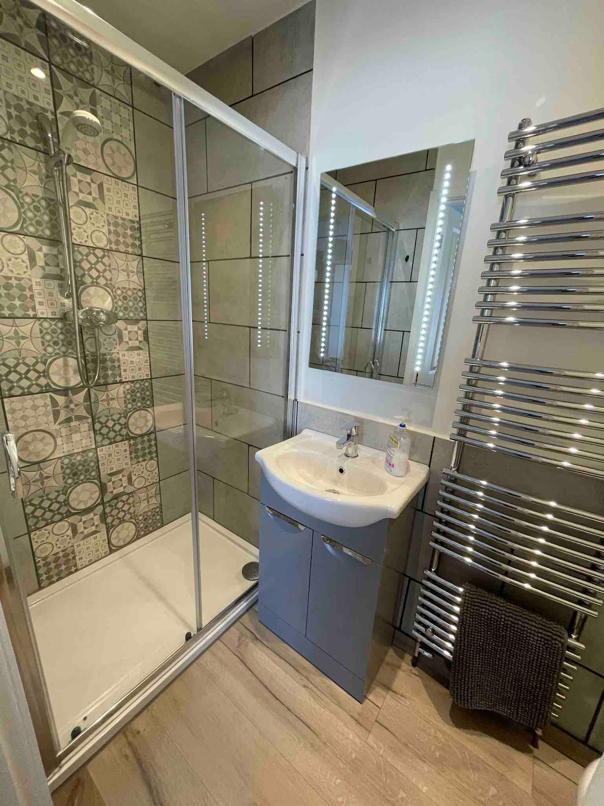 A modern bathroom features a glass-enclosed shower with unique patterned tiles and a sleek sink with storage. Illuminated mirrors reflect light, enhancing the space. A stainless steel heated towel rack displays a soft black towel, contributing to the overall contemporary design.