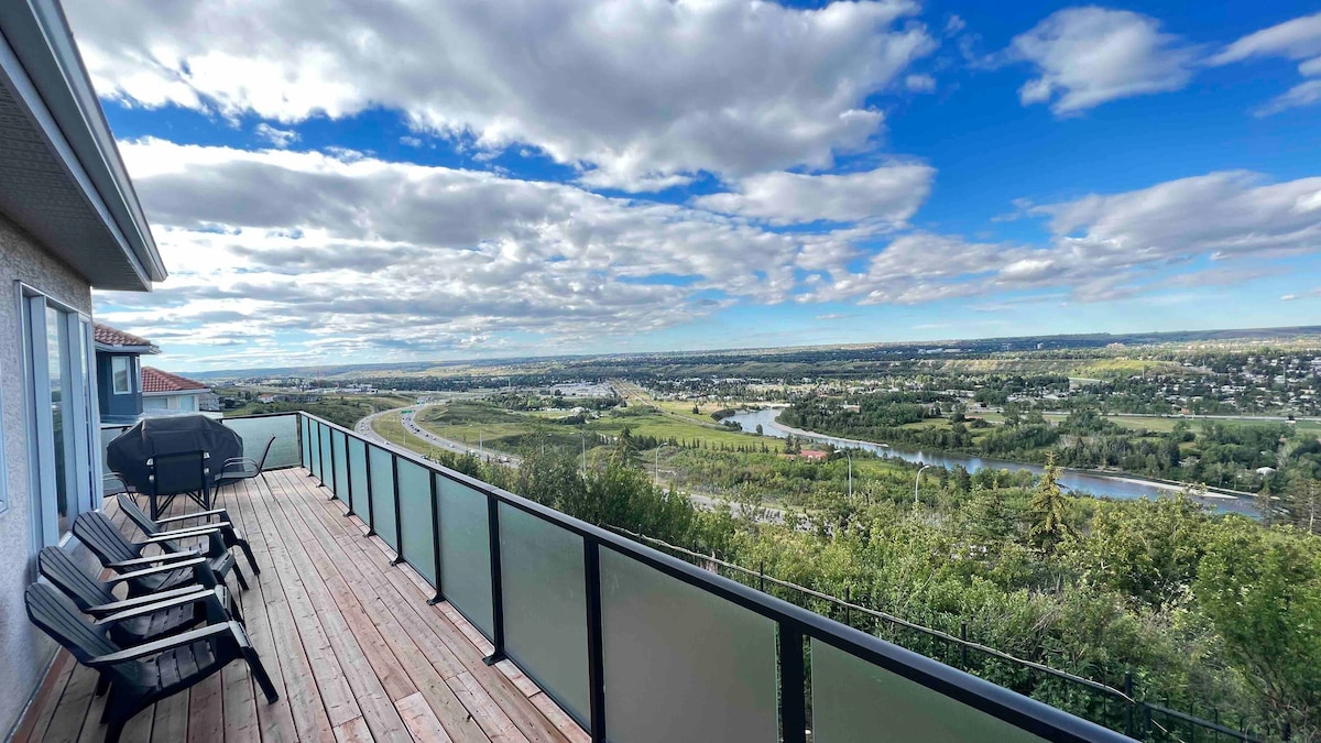 A spacious patio is lined with several outdoor chairs, providing a comfortable area for relaxation. Breathtaking panoramic views of the surrounding landscape and city are visible, with lush greenery and winding rivers adding to the serene backdrop against a partly cloudy sky.
