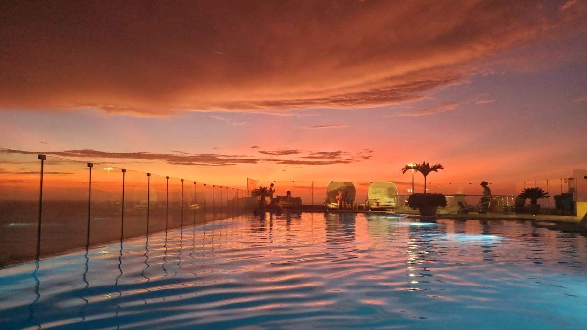 A panoramic pool is framed by a glass railing, with a vibrant sunset casting rich hues across the sky. The water reflects the colors, creating an inviting atmosphere. Palm trees and comfortable lounging areas are visible in the background.