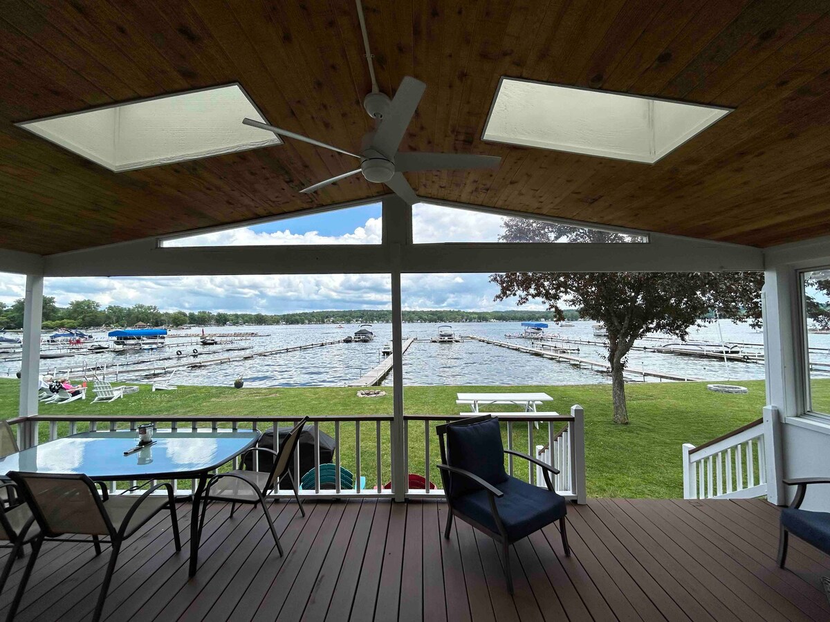 A spacious porch deck features a dining table and comfortable seating, offering a view of the lake and nearby boat docks below. Large skylights and a ceiling fan add to the airy ambiance. Green grass in the yard frames the serene water scene.