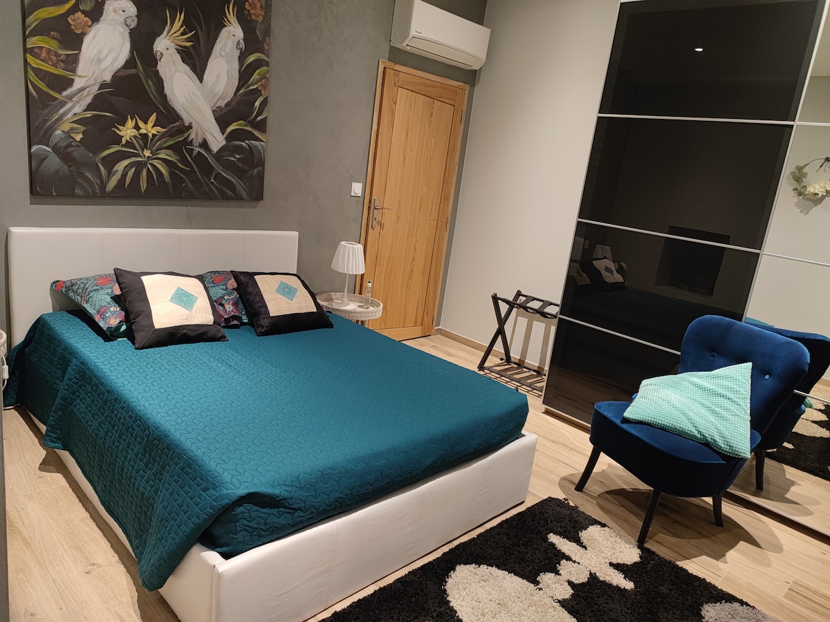 A spacious sleeping area is furnished with a large bed covered in a teal quilt and accented by decorative pillows. A modern armchair sits beside a small table, while a sleek wardrobe features mirrored panels, enhancing the room's light and openness.