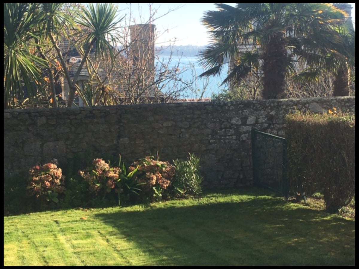 A well-maintained garden features lush green grass and vibrant flowering plants. A stone wall encloses the space, with a gate providing access. Palm trees are visible in the background, and a hint of water can be seen beyond the garden, suggesting proximity to the coast.