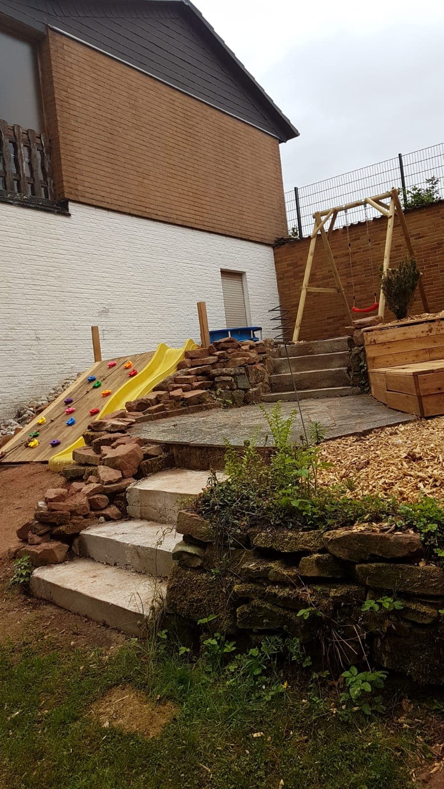 A children's play area is visible with a wooden climbing wall leading to a slide. Steps made of stone offer access to different levels, while a swing set is positioned to the right. Natural materials, including stones and wood chips, are present throughout the space.