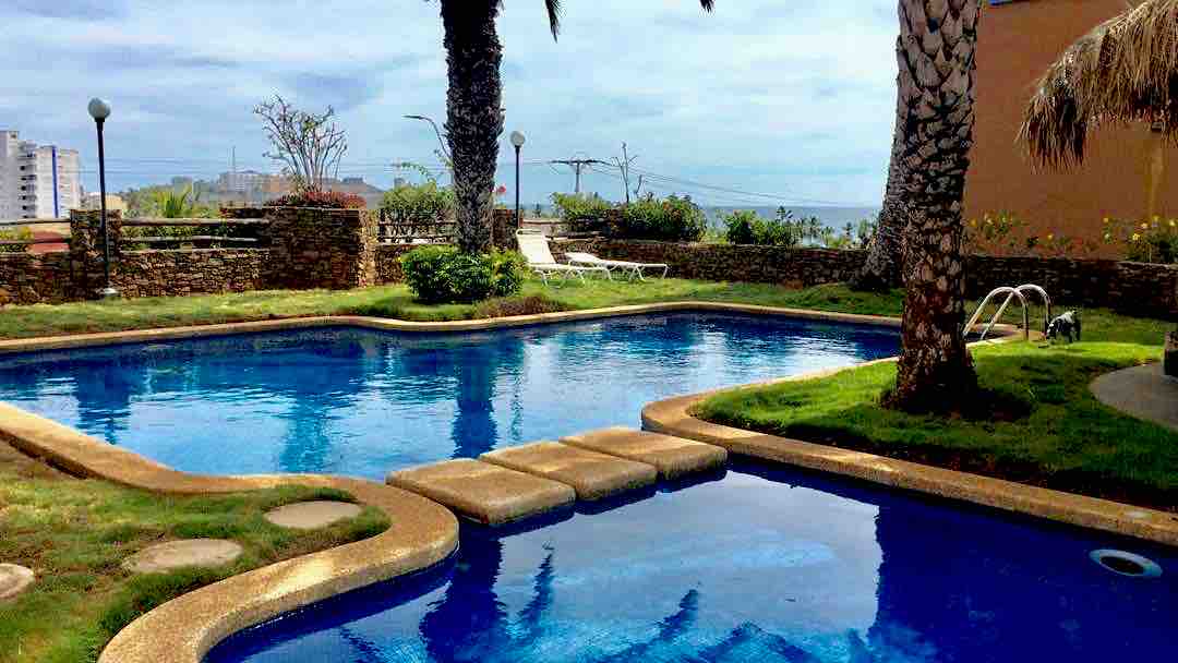 A tranquil swimming pool is framed by palm trees, with inviting blue water reflecting the sky. Lounge chairs are positioned nearby on a grassy area, and a pathway leads from the pool to the landscaped garden, creating a relaxing outdoor space.