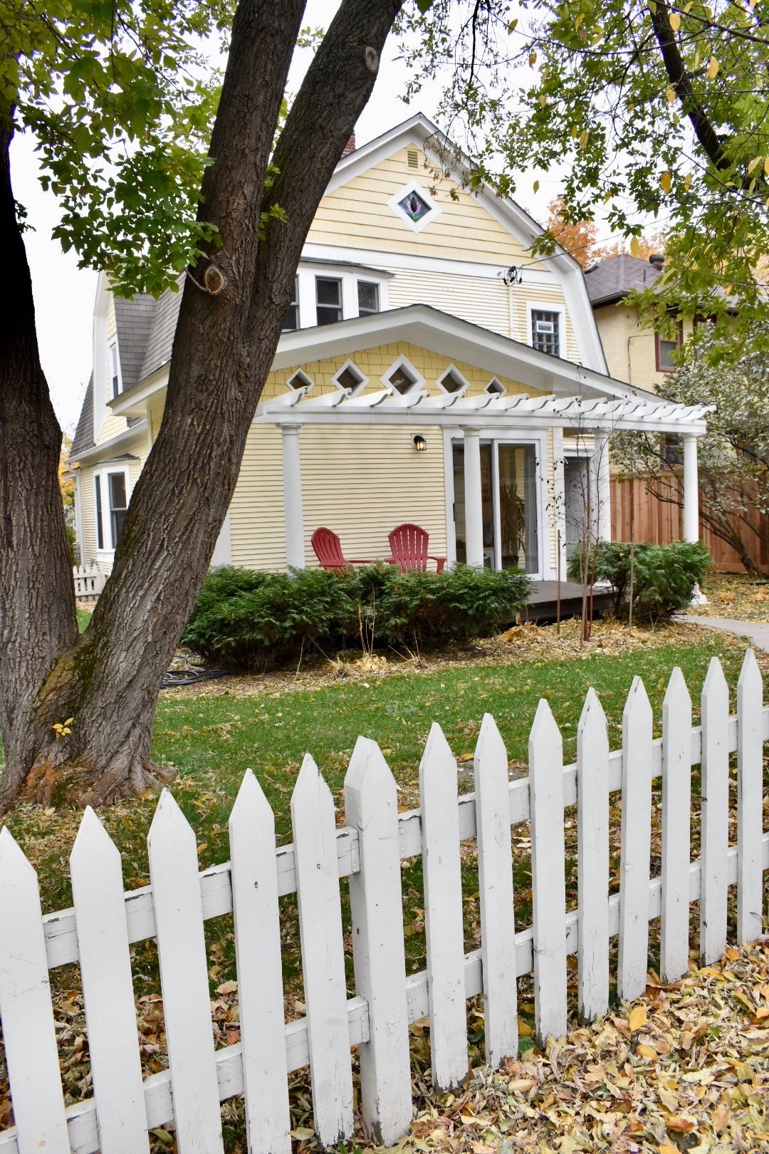 The exterior of a charming yellow house is framed by two large trees and a white picket fence. Two red chairs are visible on the porch, overlooking a manicured yard surrounded by greenery. A warm, inviting atmosphere is complemented by the architectural details of the home.