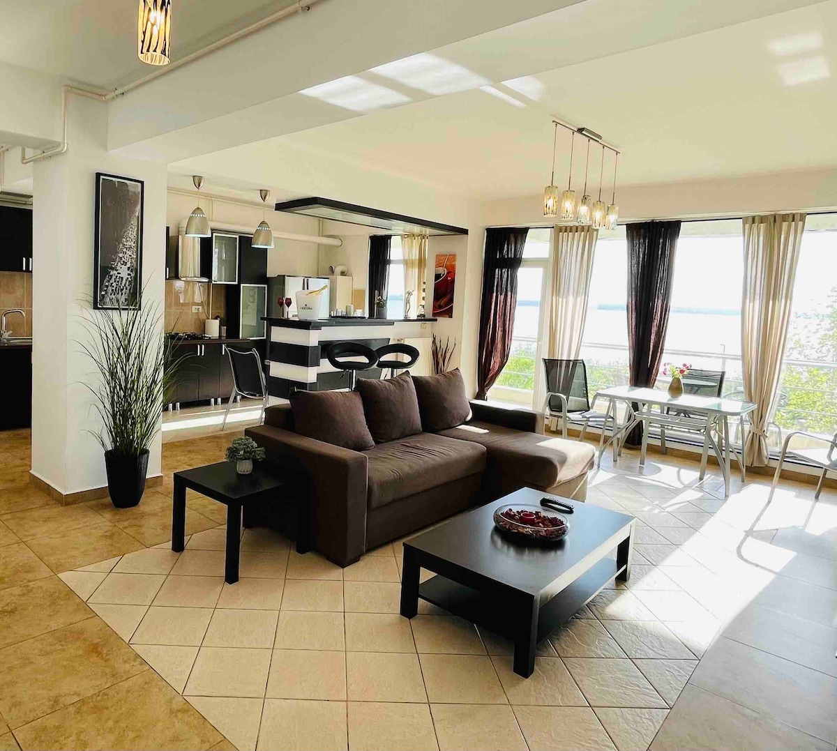 An open living area features a large sofa grouped with a coffee table, set against floor-to-ceiling windows that provide views of the lake. The space includes a dining table and a kitchen area, with natural light illuminating the modern decor.