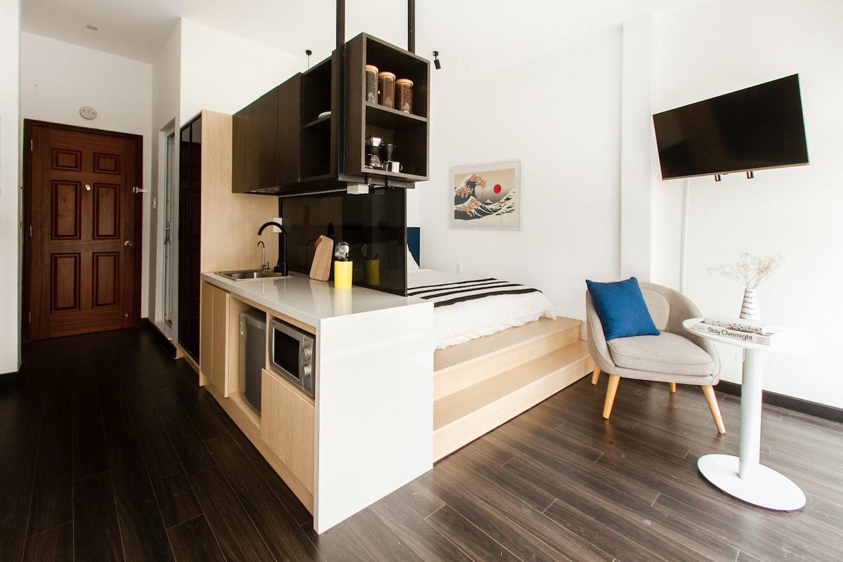 A minimalist studio is displayed with a polished wooden floor, featuring a platform bed and a compact kitchen area. A television is mounted on the wall, and a small dining table is positioned beside a plush armchair, creating a comfortable living space.