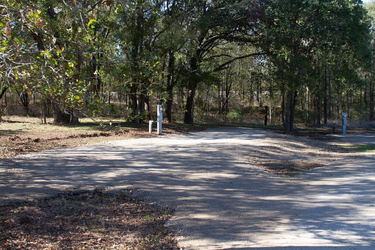A gravel pull-in site is visible, surrounded by trees, creating a natural setting. Power and water hookups are positioned along the site. The ground is mostly clear of leaves, providing a spacious area for parking RVs or tents.