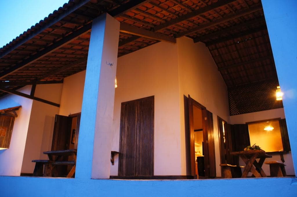 A spacious outdoor area is framed by sturdy columns, showcasing an inviting entryway. Wooden doors open to reveal an interior space, while warm lighting fixtures hang from the ceiling. Natural materials are incorporated throughout, creating a harmonious blend of indoor and outdoor elements.