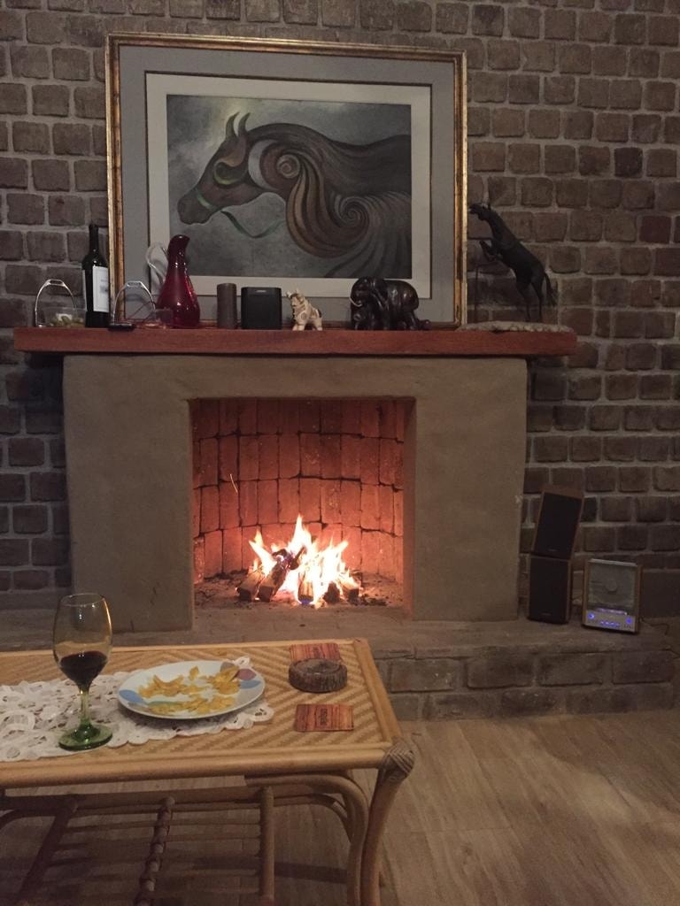A rustic fireplace with a warm flame is set against a textured stone wall. A glass of red wine and a plate of snacks are arranged on a woven coffee table, with decorative items and a framed artwork visible on the mantel.