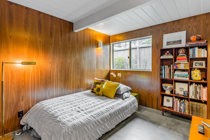 - Snuggle up in this stylish nook with rich wood paneling and bright natural light
- Thoughtful touches like plush pillows and a soft gray duvet create cozy vibes
- Browse your favorite books from the vibrant shelf packed with character

