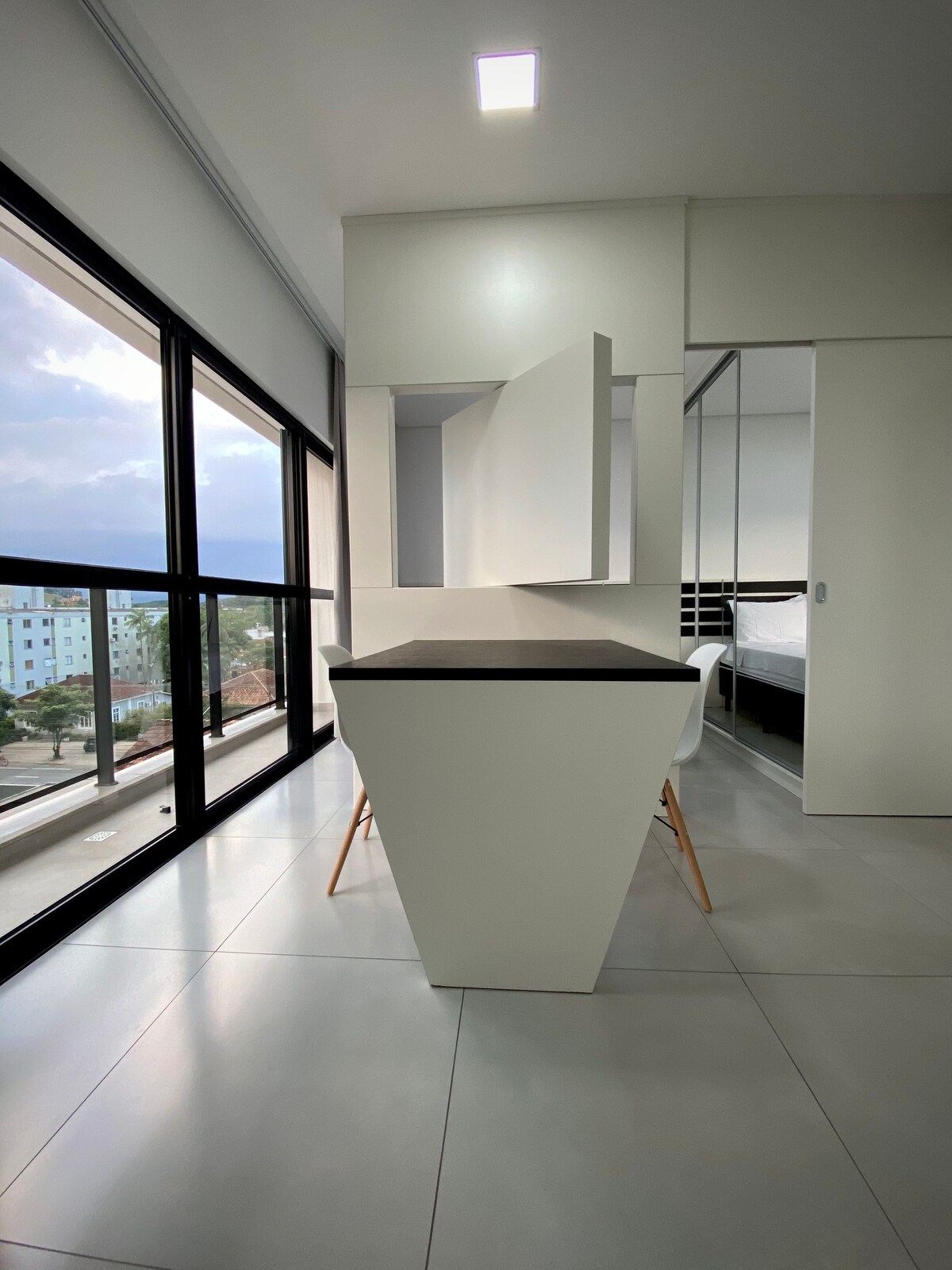 A modern workspace is featured in the foreground, complemented by a geometric table and white chairs. Expansive windows allow natural light to flood the space, providing a view of the city skyline. Minimalist cabinetry adds functionality to the room.