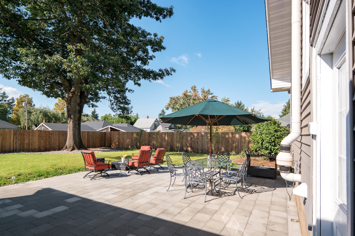 An outdoor patio area is depicted, featuring several lounge chairs and a dining table sheltered by a green umbrella. A spacious lawn is visible beyond the paved surface, enclosed by a wooden fence and shaded by a large tree.
