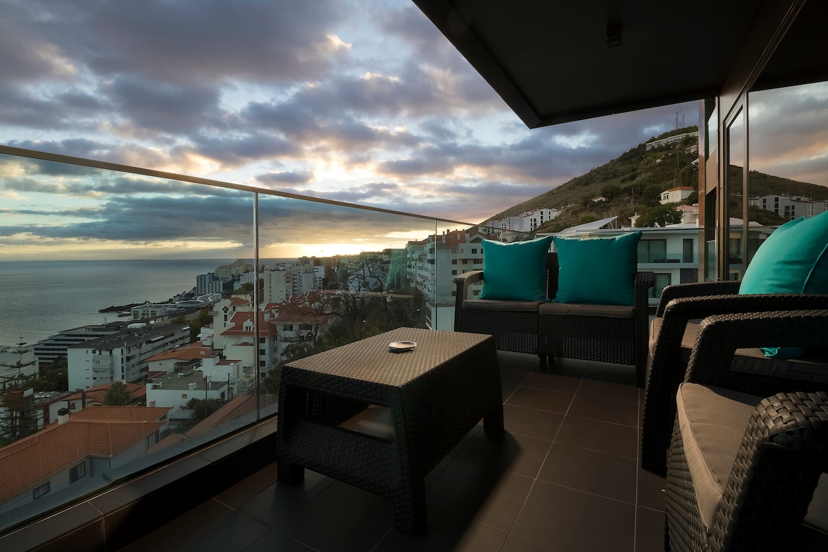 A spacious balcony is presented, featuring a comfortable seating area with two armchairs and a coffee table. A breathtaking view of the coastline and distant mountains is visible, enhanced by a dramatic sky at sunset.