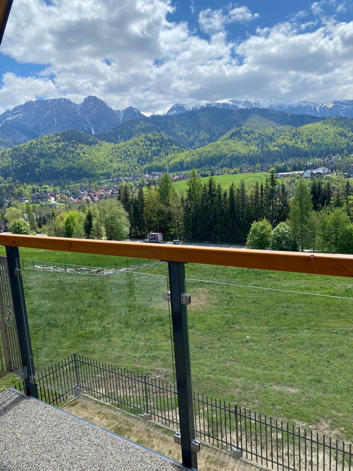 A panoramic view of lush green hills and majestic mountains is framed by a glass balcony railing. The landscape features a mix of forest and grassy fields, with patches of snow visible on the mountain peaks under a bright blue sky with scattered clouds.