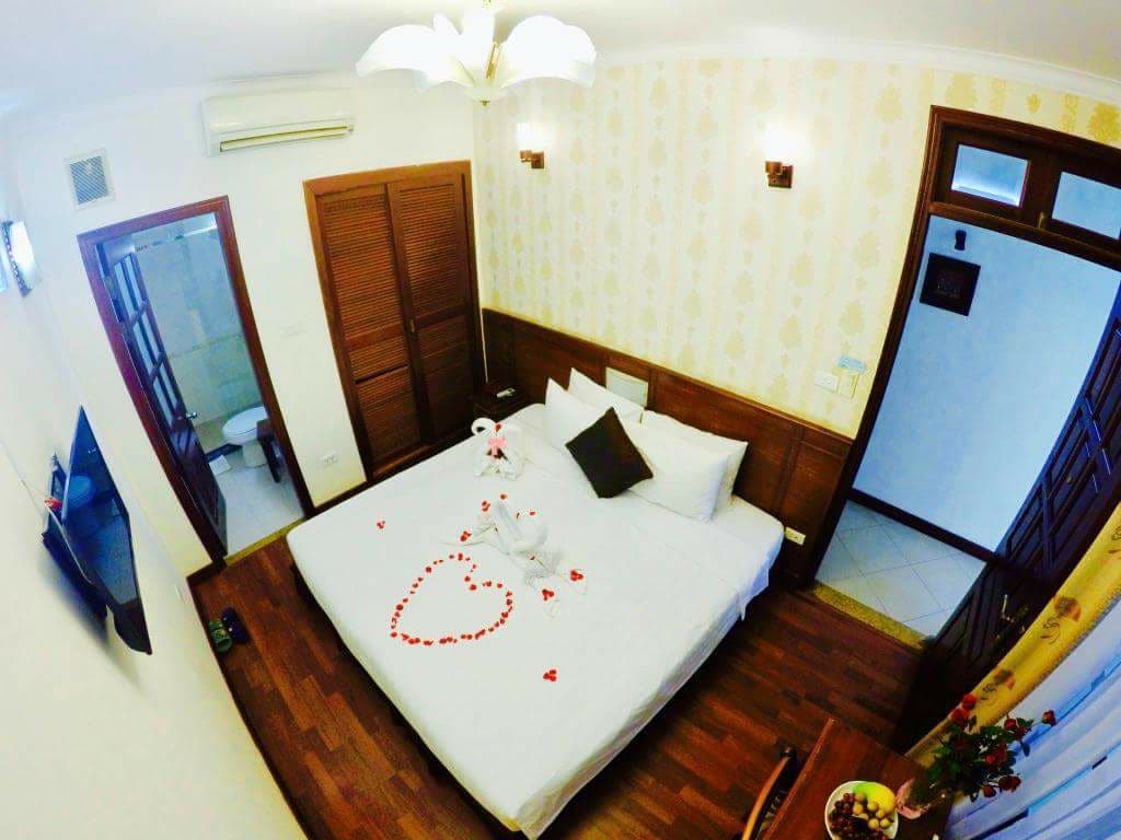 An aerial view of a well-appointed bedroom features a spacious bed adorned with decorative towels arranged in a heart shape, surrounded by scattered rose petals. Natural wood accents complement the light-colored wallpaper. A television mounted on the wall is visible, along with an en-suite bathroom door.