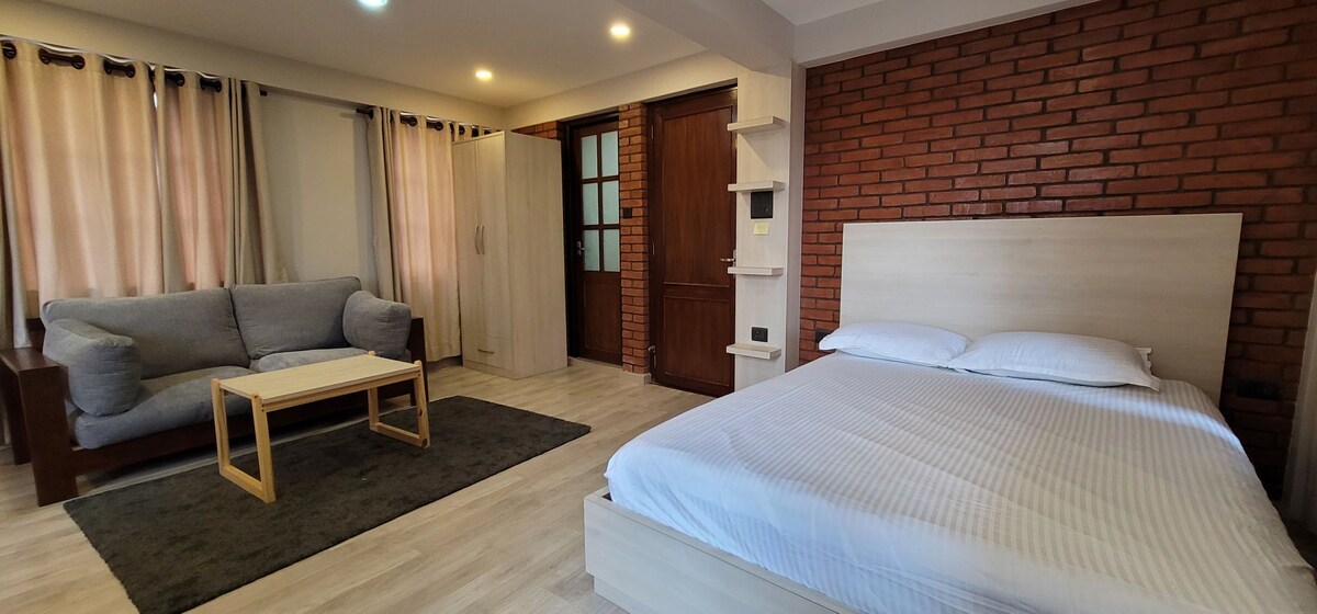 An inviting living space showcases a spacious bed adjacent to a comfortable sofa and a wooden coffee table. Natural light filters through sheer curtains, and a brick wall adds texture to the room. A door leads to the entrance, providing access to the outside.
