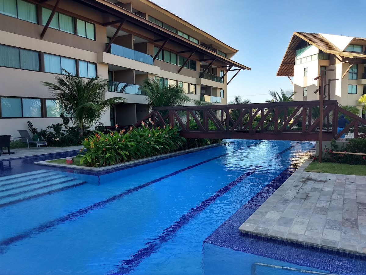 The image captures a serene pool area within a residential complex. A wooden bridge spans over the blue water, surrounded by tropical plants. The three-story buildings in the background feature balconies, with palm trees enhancing the tropical ambiance.
