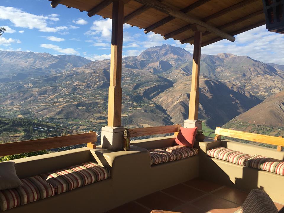 A shaded outdoor seating area features built-in benches with striped cushions, offering views of the expansive mountainous landscape. The sky is clear with scattered clouds, and the terrain below displays a patchwork of fields and valleys.