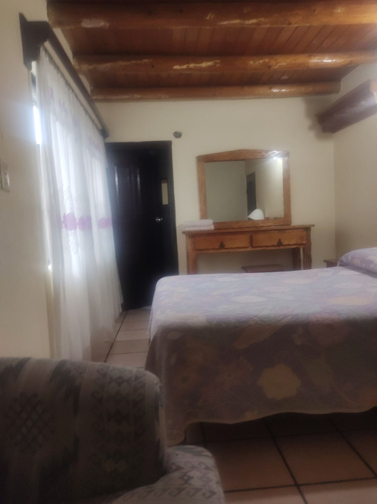 A cozy bedroom is highlighted by a spacious bed covered with a patterned blanket. Natural light enters through sheer curtains, enhancing the room's brightness. A wooden dresser is positioned against the wall, while a comfortable armchair is placed nearby, complemented by tiled flooring.