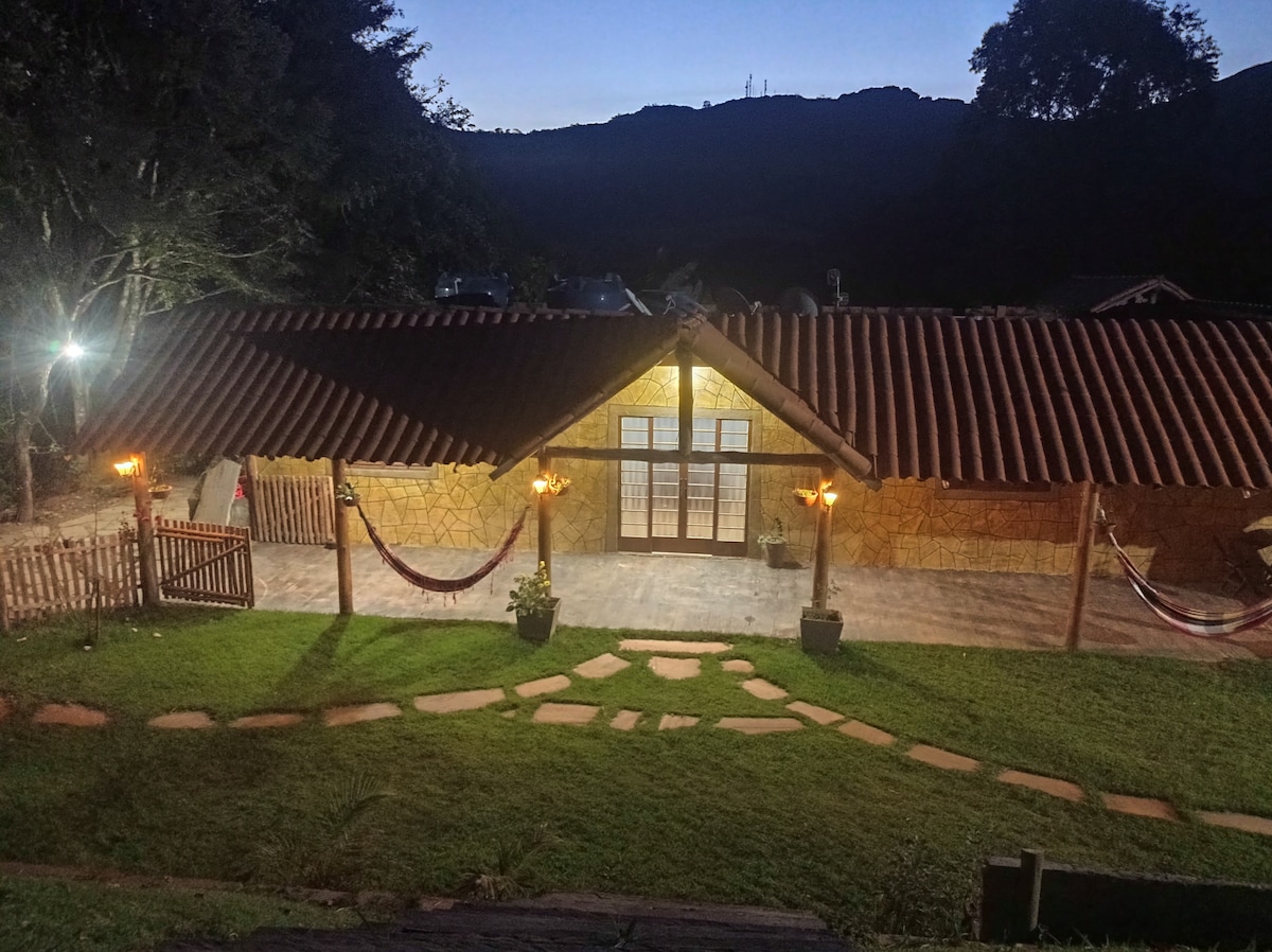 The exterior of the rustic house is shown at dusk, with warm lighting illuminating the entryway. A stone path leads to the main entrance, flanked by decorative plants. Hammocks are suspended in the yard, and the surrounding landscape features lush greenery and distant hills.