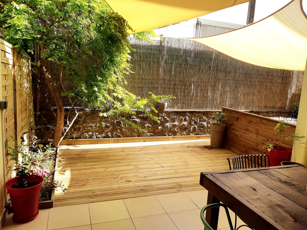 A peaceful outdoor space features wooden decking, complemented by potted plants and a shaded dining table. Natural light filters through a canopy, enhancing the greenery from a nearby tree. A decorative stone wall provides a serene backdrop, promoting a relaxing atmosphere.
