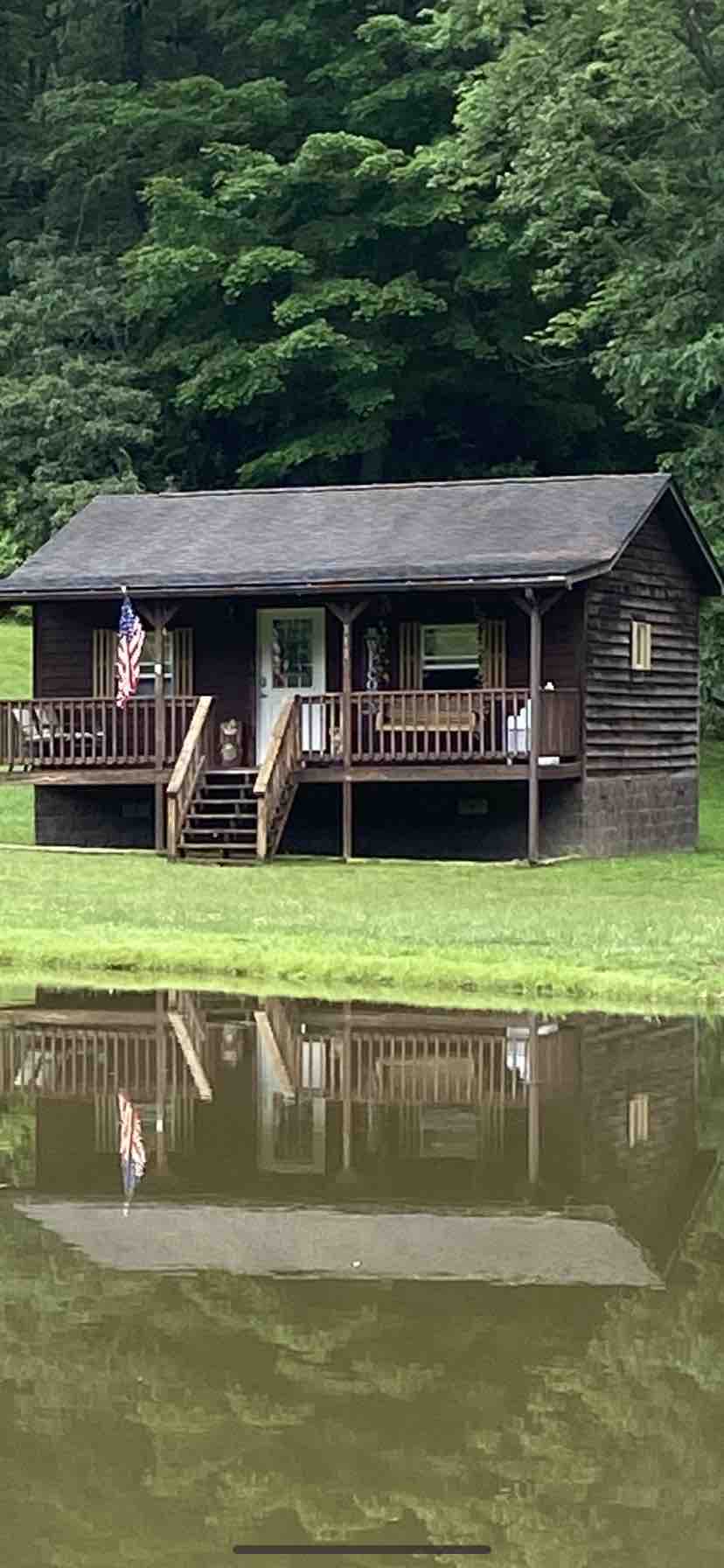 A rustic cabin is set beside a fishing pond, surrounded by lush greenery. The wooden structure features a front porch with steps leading down to the water's edge, where reflections create a serene atmosphere. An American flag is displayed, contributing to the classic country aesthetic.