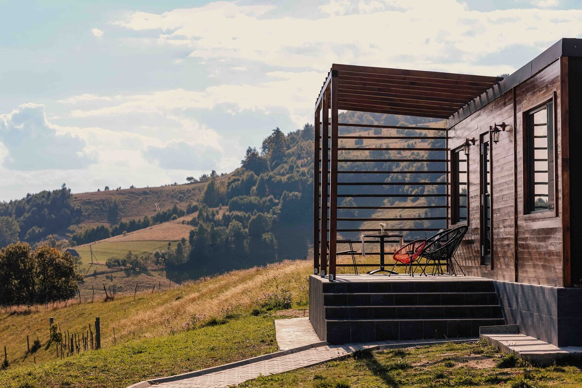 The outdoor area features a wooden deck with modern seating arranged for relaxation. Expansive views of rolling hills and trees are visible in the background, with soft clouds scattered across the sky.