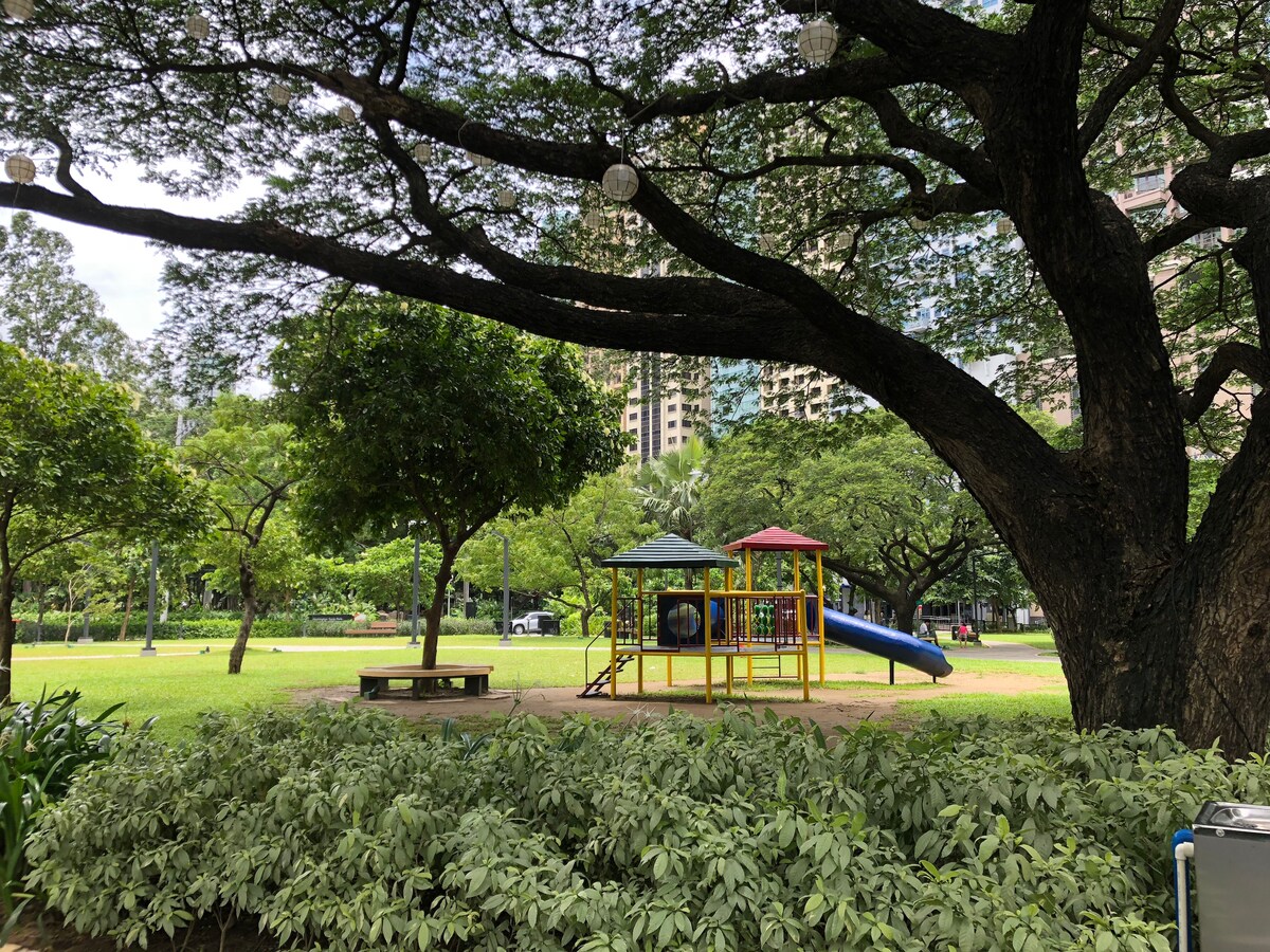 A colorful playground is featured within a grassy area, surrounded by lush green trees. A blue slide and two red-and-green canopies adorn the play structure. Nearby, a wooden bench is positioned under the shade of a large tree, providing a serene spot for relaxation.