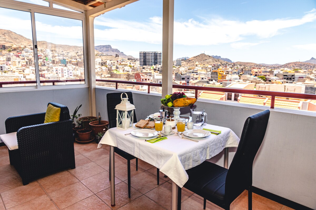 An outdoor dining area features a table set for breakfast, including dishes, beverages, and fresh fruit. Two chairs and a black wicker sofa are positioned nearby. The expansive view displays the surrounding cityscape and hills, with a lantern and potted plants enhancing the setting.