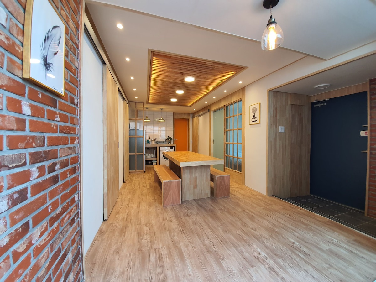An open living area features a long wooden dining table surrounded by ample space. Exposed brick walls contrast with light wood finishes above the table. Large windows allow natural light to fill the room, enhancing the cozy atmosphere leading to the kitchen area.
