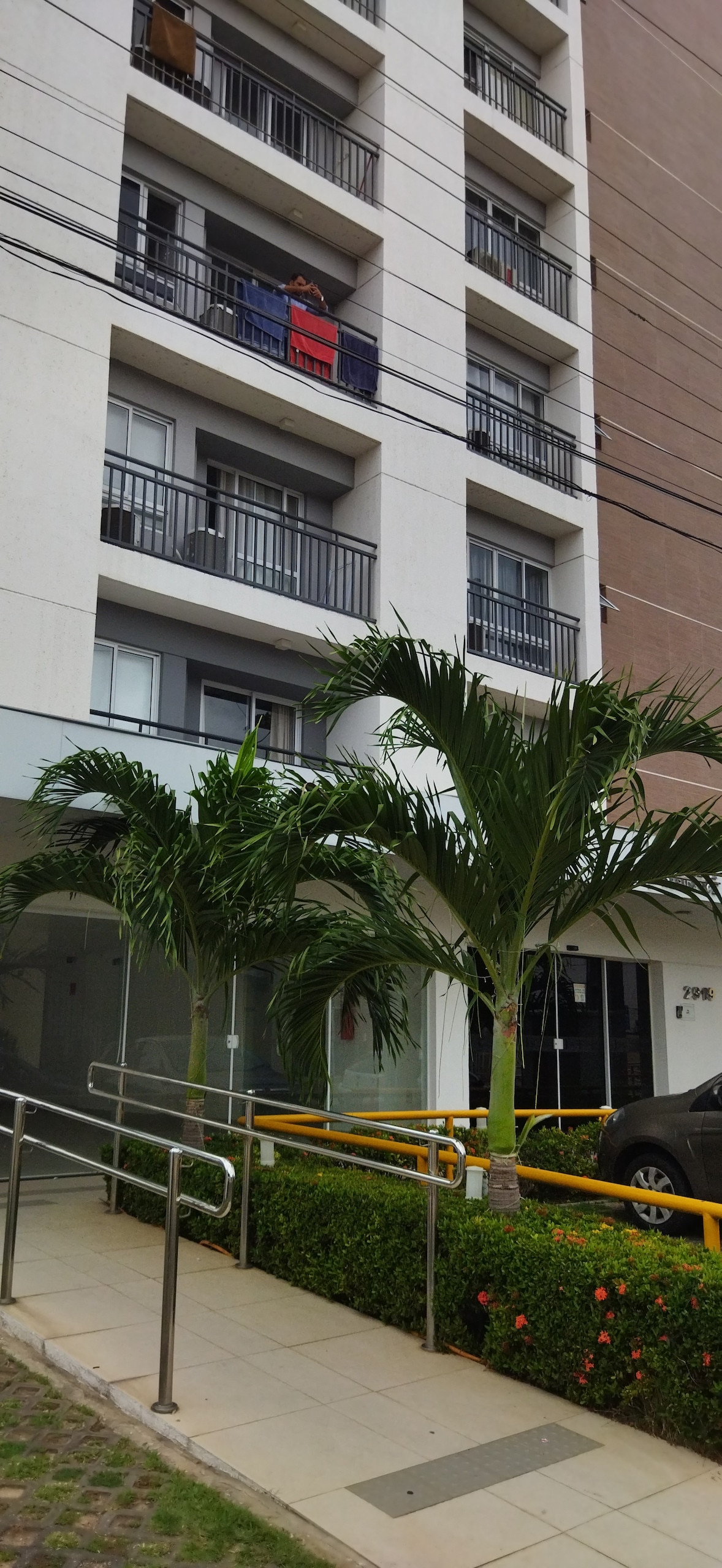 The exterior of a multi-story building is displayed, featuring balconies with railings on each floor. Lush palm trees frame the entrance with a pathway leading to the door. A vehicle is parked nearby, and the surrounding area is well-maintained.