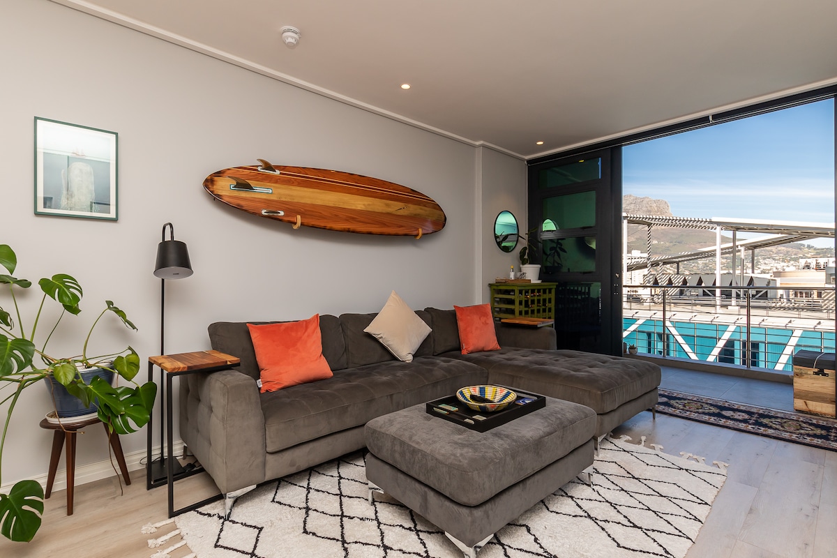 A spacious living area features a sectional sofa with orange accents and a large ottoman. A surfboard is mounted on the wall, and a plant adds a touch of greenery. Large glass doors open to reveal views of the city and mountains.