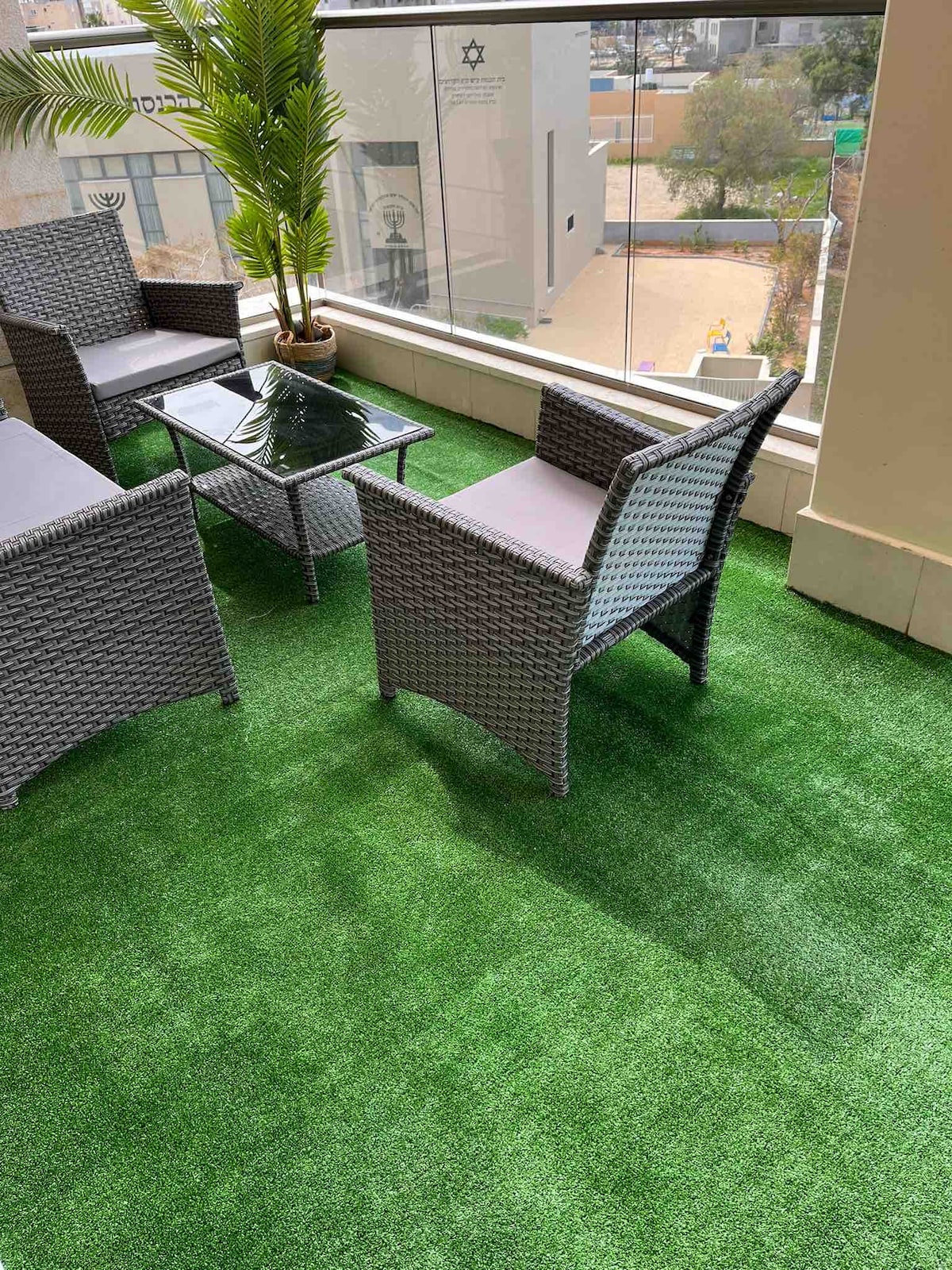 A cozy seating area is situated on a balcony, featuring a glass-topped table surrounded by four woven chairs. The floor is covered in artificial grass, complemented by a decorative potted plant nearby, offering a relaxed outdoor atmosphere.