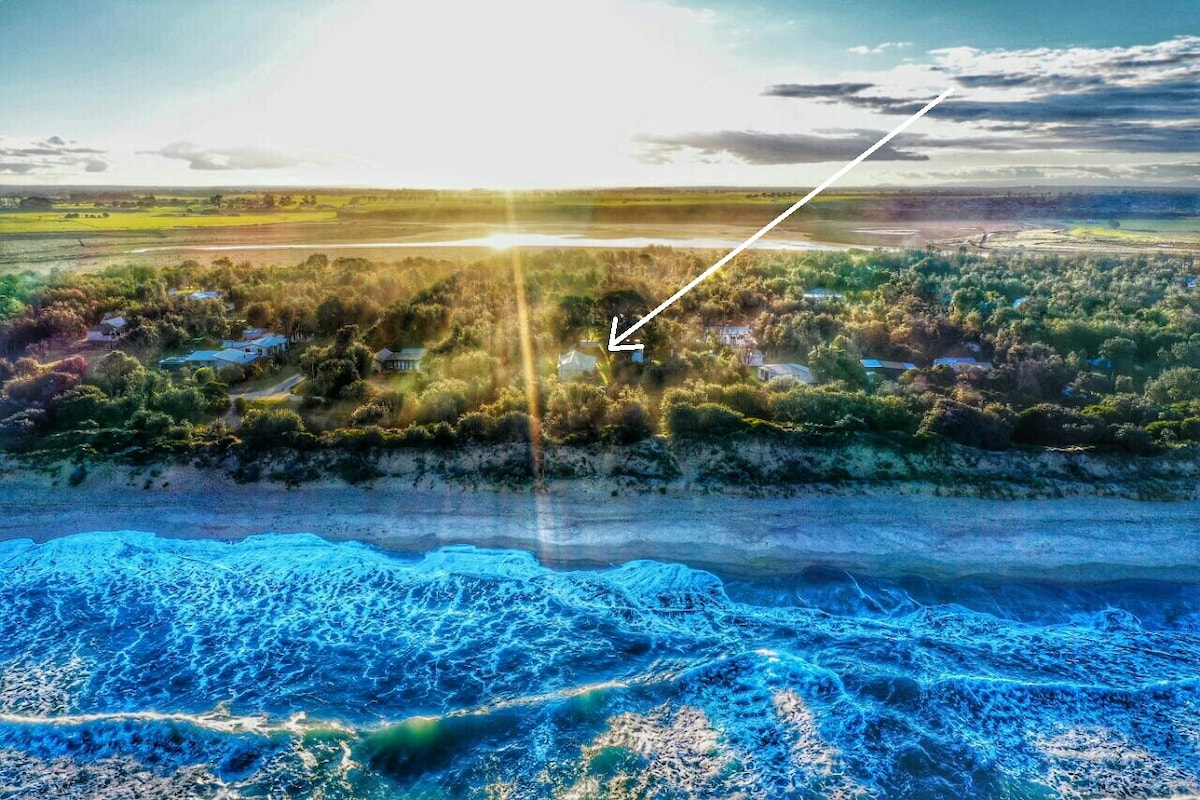 An aerial view captures the beachfront location of the house, with the vibrant blue waves gently lapping at the shore. Soft sunlight bathes the landscape, highlighting the natural surroundings and creating a serene atmosphere. The house is positioned among trees, offering a tranquil coastal retreat.