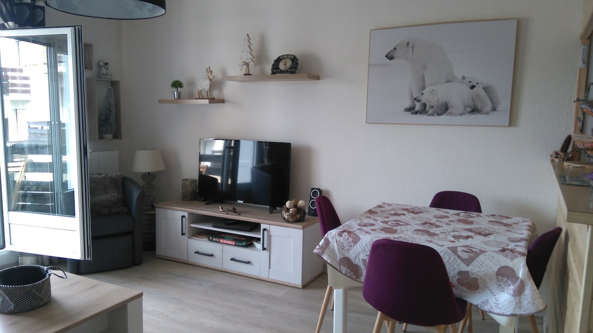 A cozy living area features a flat-screen TV on a light wooden console, paired with a round dining table and purple chairs. Decorative elements, including a polar bear print, are displayed on the wall and shelves. A window allows natural light to enter the space.