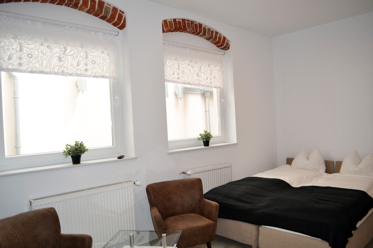 A well-lit sleeping area features a spacious bed with white linens, flanked by two windows dressed with sheer lace curtains. Two armchairs are positioned beside a glass table, complementing the minimalist decor. Exposed brick accents can be seen on the walls, adding a touch of warmth.
