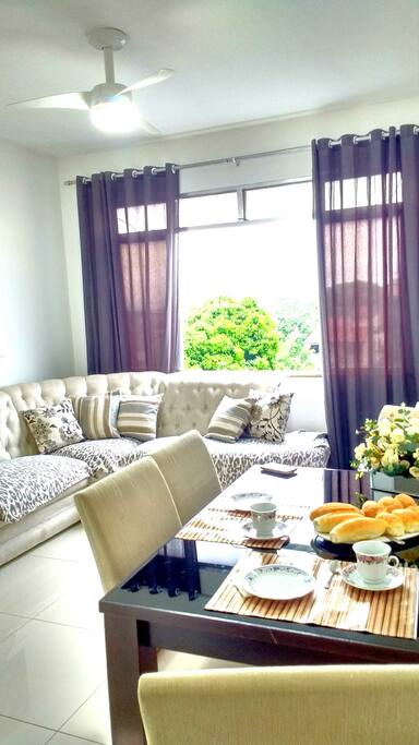 Sala com sofá para cinco pessoas, mesa para quatro pessoas, painel com TV a cabo, ventilador de teto e uma janela grande.