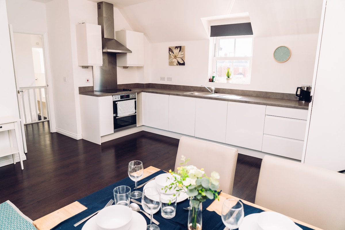 A modern kitchen space features sleek white cabinetry and a full kitchen setup, including an oven and hob. A dining area is visible with a neatly set table, showcasing plates and glassware, and a small decorative arrangement of greenery.