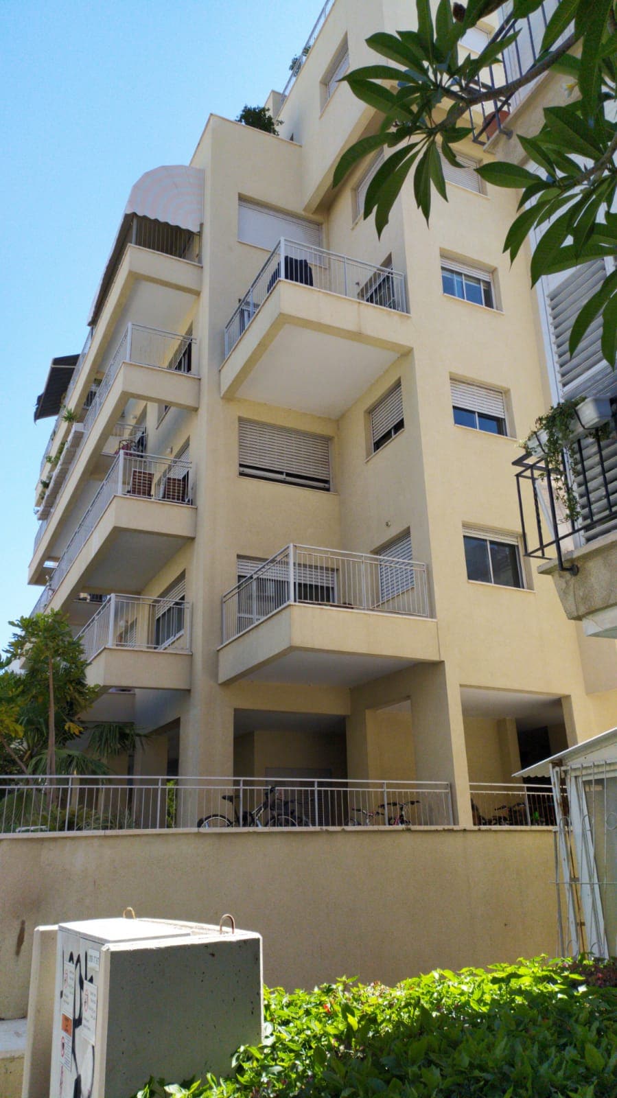 The modern apartment building features multiple balconies and a light-coloured façade. The ground level is surrounded by greenery, and bicycles can be seen secured near the entrance. Sunlight casts shadows, highlighting the contemporary structure against a clear blue sky.