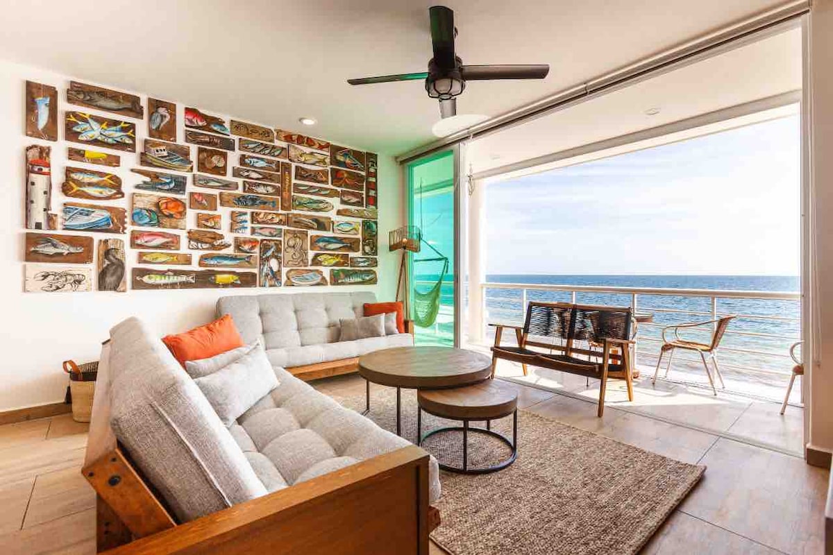 A spacious living area features a cozy seating arrangement with a large sectional sofa and wooden lounge chairs. A vibrant artwork decorates the wall, and large glass doors provide an unobstructed view of the ocean. Natural light fills the space, enhancing the coastal ambiance.