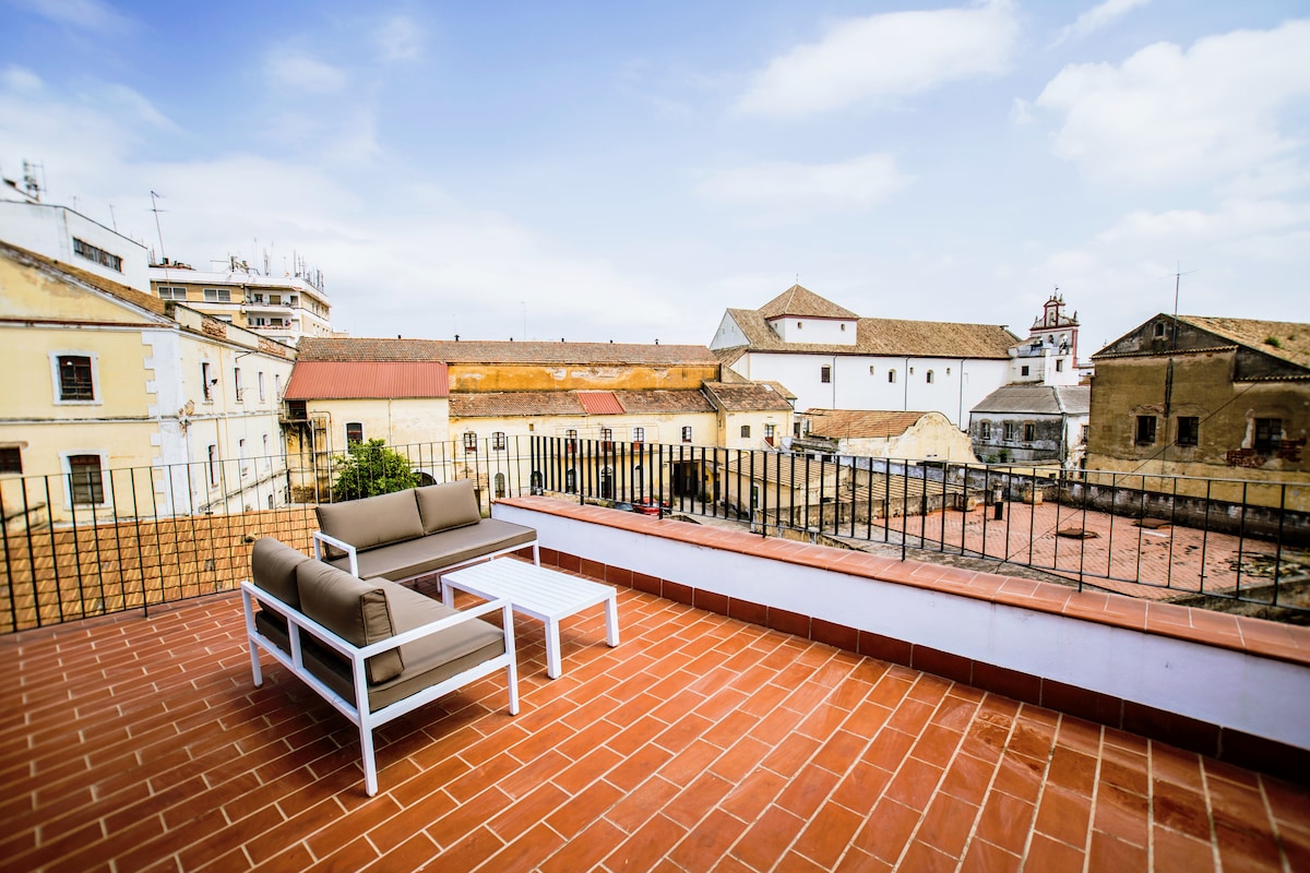 A private terrace offers a spacious seating area furnished with comfortable sofas and a small table. Surrounding views include historical buildings and rooftops, presenting a blend of architecture under a clear sky.
