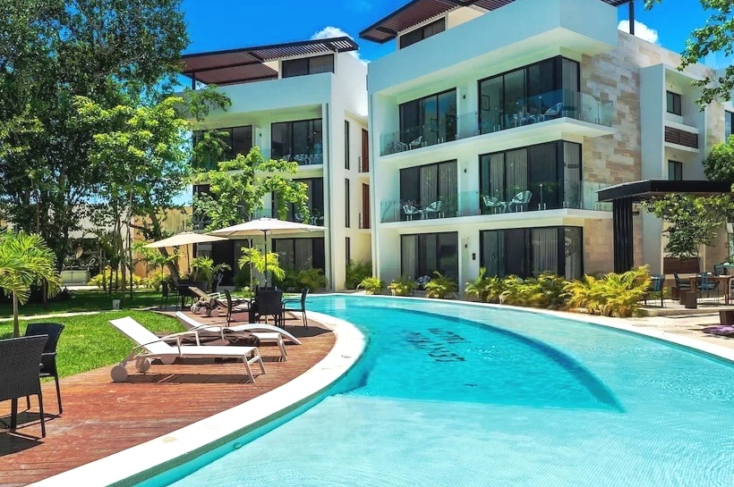 A modern three-story building is shown, featuring large glass doors and balconies. Surrounding the structure is a curved swimming pool, bordered by lush greenery and lounge chairs. Umbrellas provide shaded seating areas, enhancing the tropical ambiance of the outdoor space.
