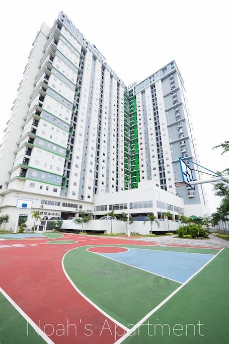 The apartment complex is viewed from the ground, featuring a tall, modern building with multiple stories. The vibrant court below showcases colorful sections for basketball and other recreational activities, surrounded by greenery and landscaping.