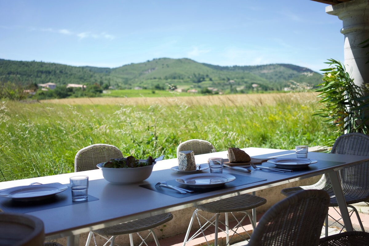 An outdoor dining area is set with a long table, surrounded by greenery and offering a view of rolling hills in the distance. Plates, glasses, and bowls are placed neatly on the table, ready for a meal in nature.