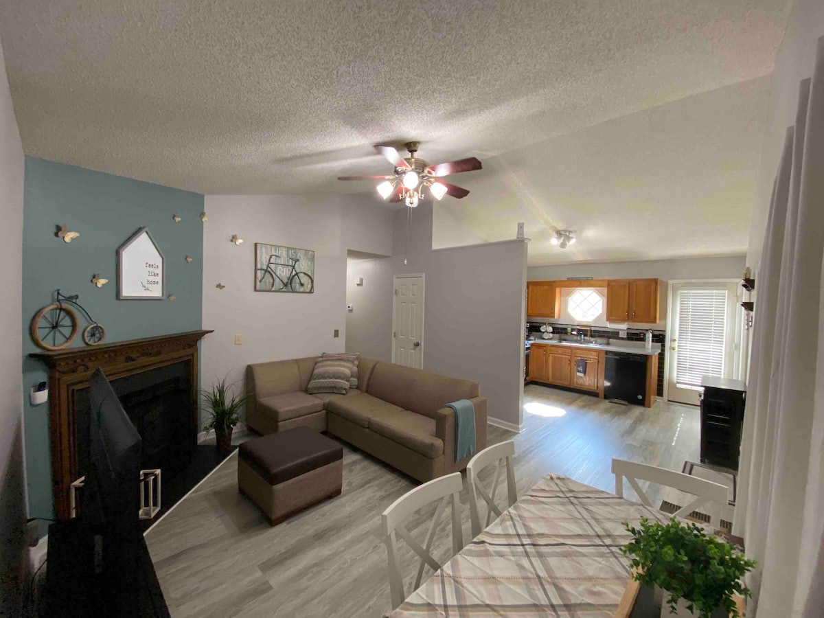 A spacious living area is showcased, featuring a cozy sectional sofa with an accompanying ottoman. A dining table is set for six, and a kitchen with wooden cabinetry is visible in the background. Natural light fills the space through large windows.