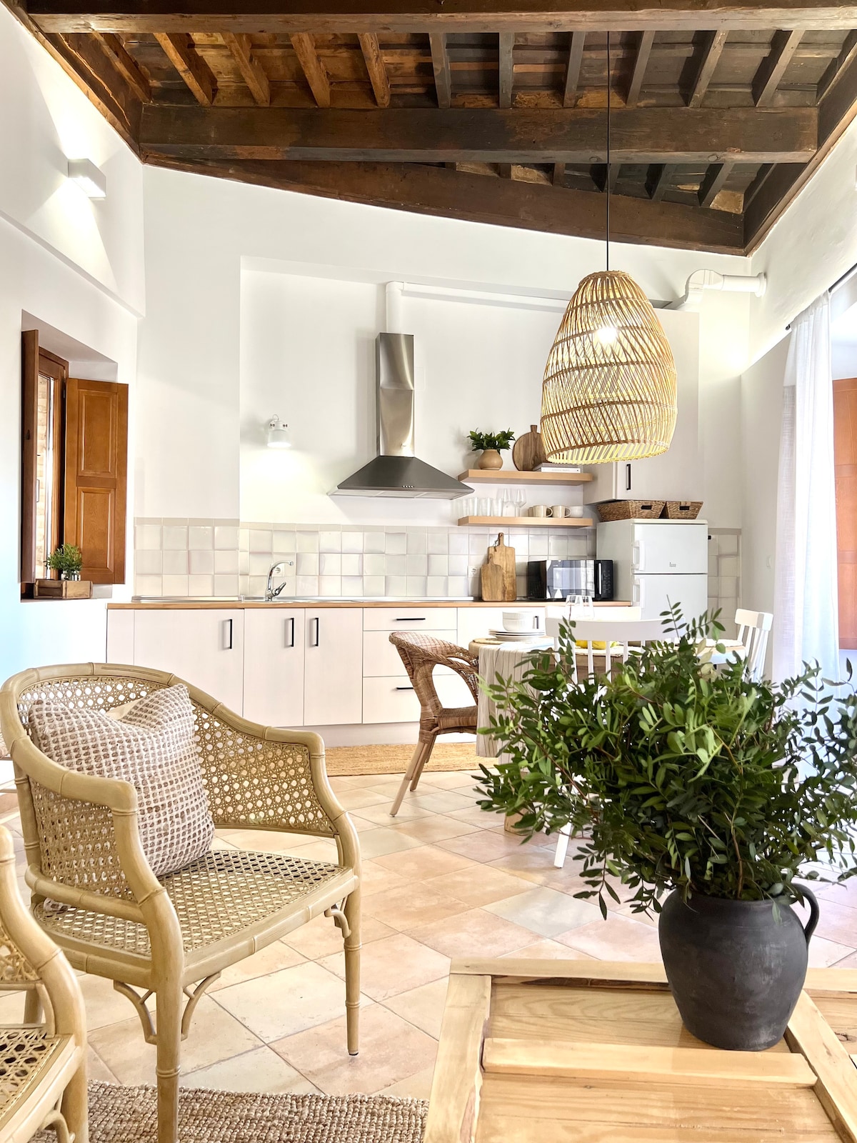 A charming kitchen area features light wood cabinetry and modern appliances, complemented by terracotta tile flooring. Natural light fills the space, highlighting a rattan pendant light and a cozy seating arrangement with woven chairs. A small table with a plant centerpiece adds a touch of greenery.