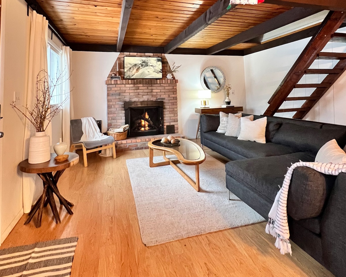 A spacious living room is showcased, featuring a brick fireplace framed by wooden beams. A comfortable couch and a stylish coffee table are arranged on light wood flooring. Natural light enters through windows, complemented by simple decor elements, creating a cozy gathering space.