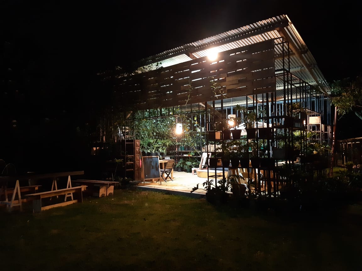 The garden area is illuminated at night, revealing a spacious structure with a metal roof. Soft lights hang from above, highlighting the surrounding greenery and potted plants. Outdoor seating areas, framed by lush vegetation, invite relaxation in a tranquil environment.